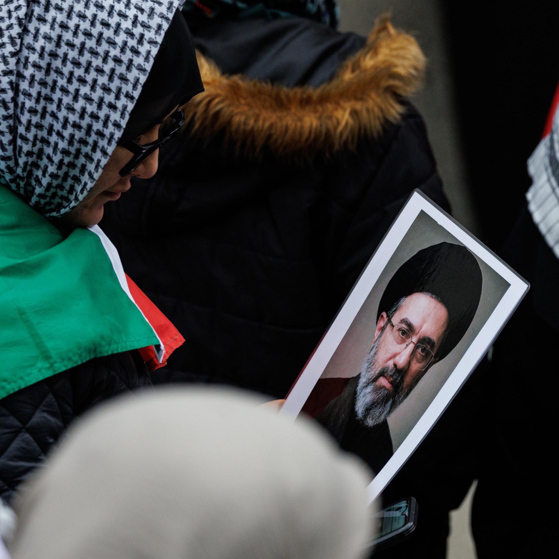 Das Bild zeigt mehrere Demonstrantinnen mit Kopftüchern in der Nahaufnahme. Eine von ihnen hält das kleine Foto in der Hand und schaut darauf. 