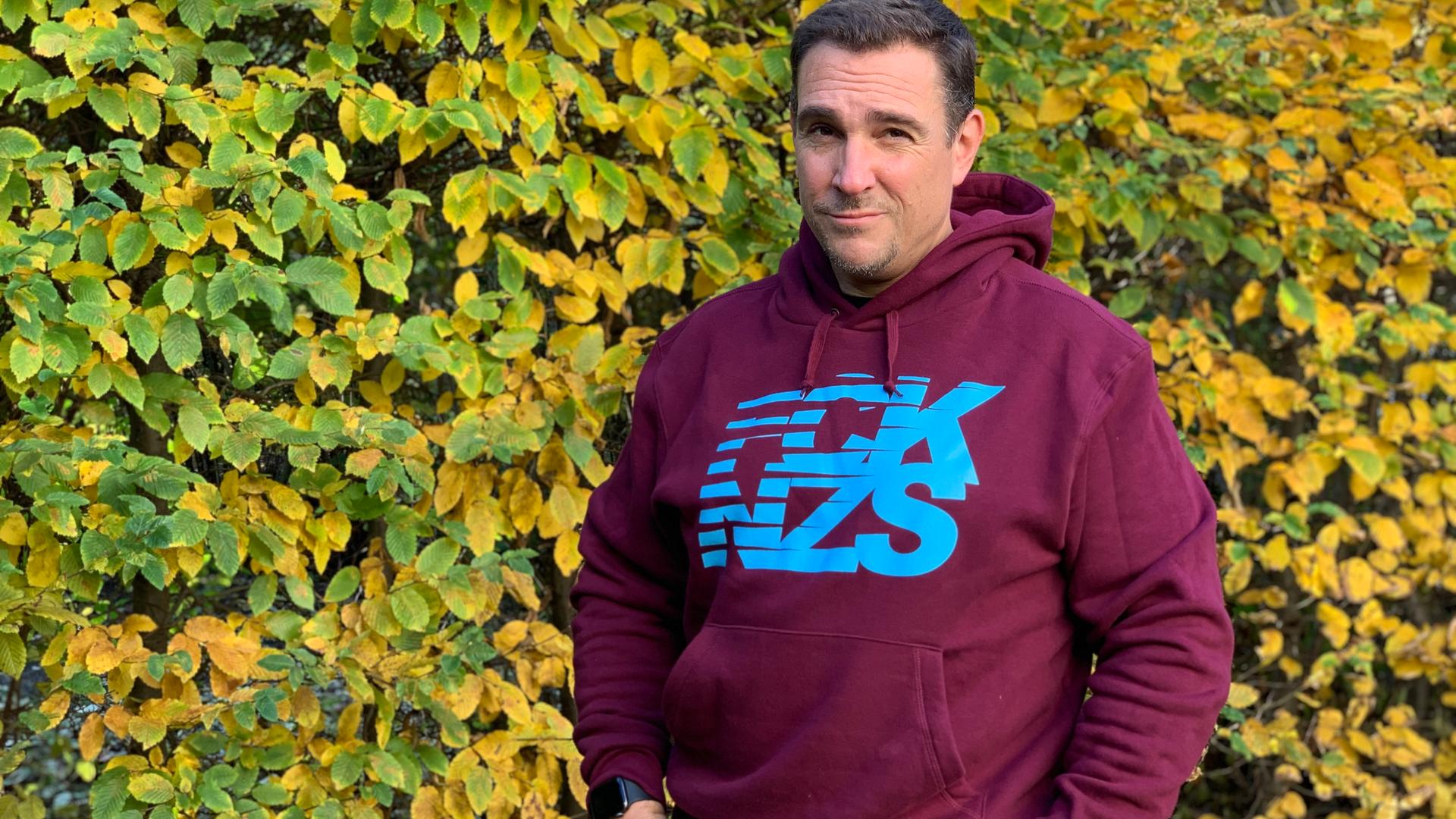 Oliver von Dobrowolski steht mit einem FCK NZS Pullover vor einer Hecke und schaut in die Kamera.