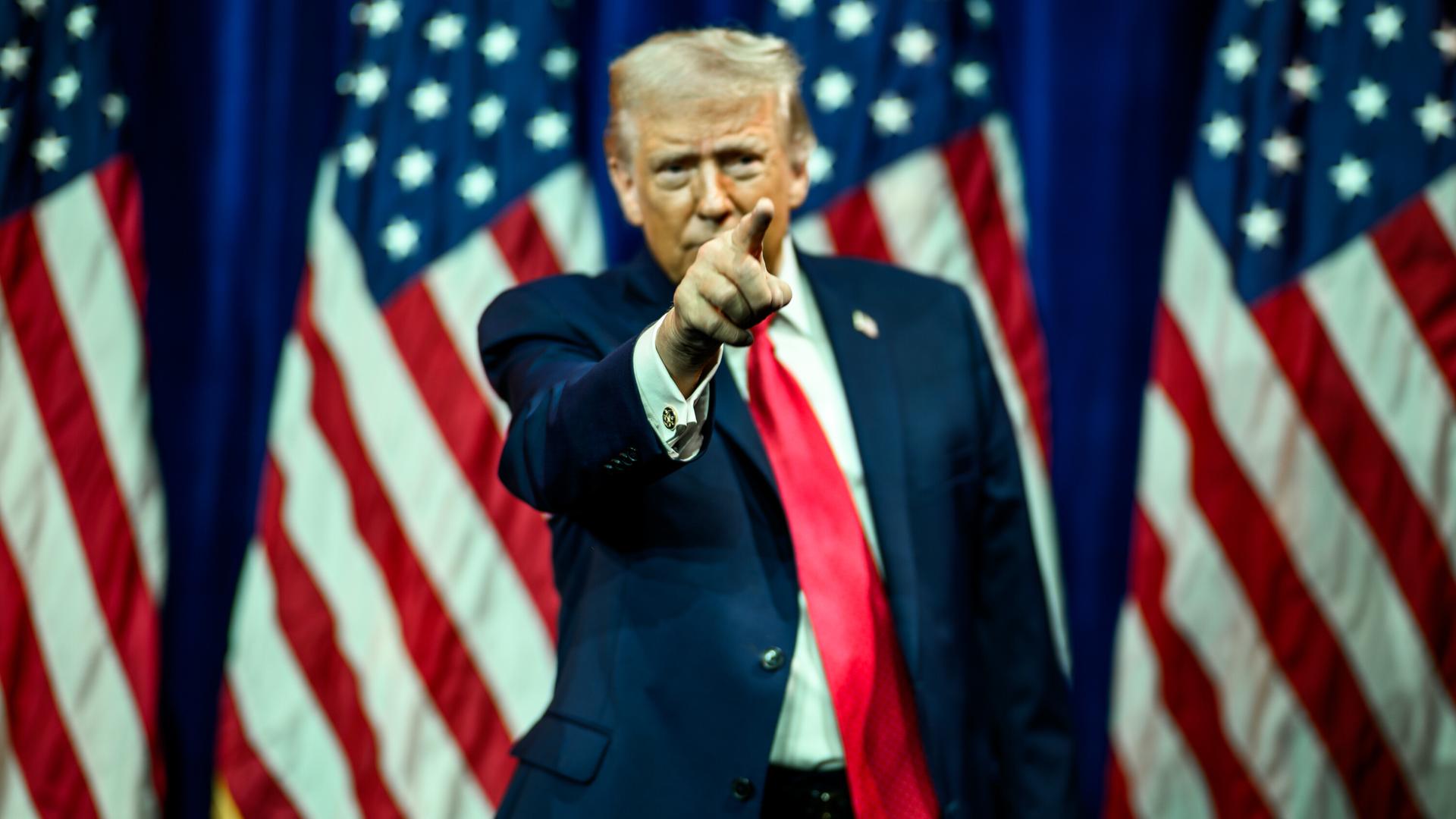 US-Präsident Donald Trump steht vor vier US-Flaggen und deutet mit dem rechten Zeigefinger auf etwas.