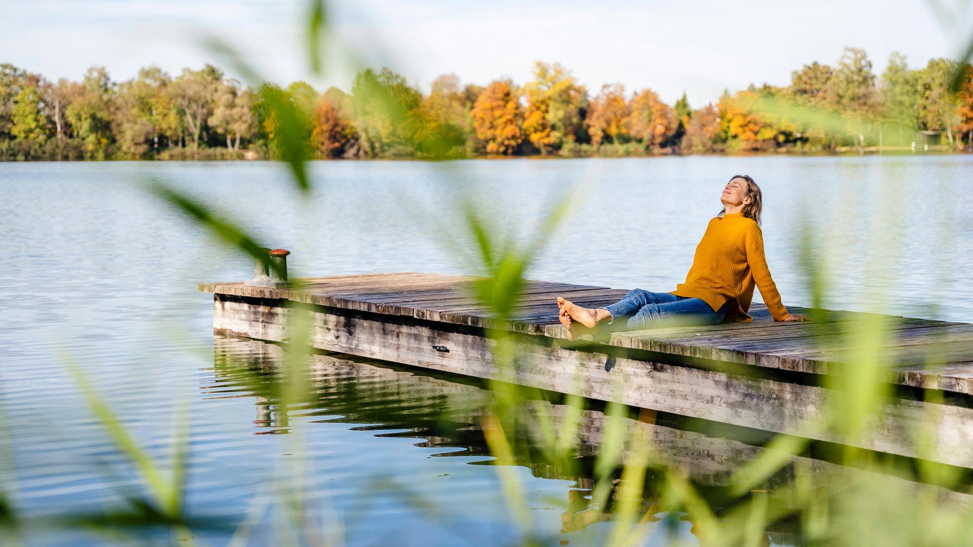 Eine Frau im gelben Pullover sitzt in entspannter Pose auf einem Holzsteg am See.