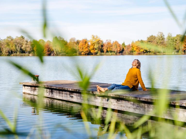 Eine Frau im gelben Pullover sitzt in entspannter Pose auf einem Holzsteg am See.