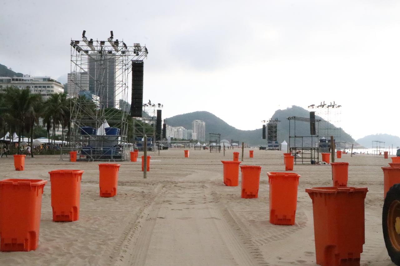 An der Copacabana in Rio De Janeiro werden die letzten Vorbereitungen für die große Silvesterparty getroffen. Eine große Bühne steht am Strand, vorne sind viele orange Mülltonnen zu sehen. 