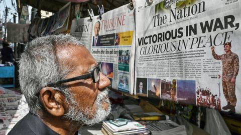 Ein Mann steht in der Stadt Karachi in Pakistan vor einem Zeitungsstand. Er liest eine Zeitung, die dort hängt. Auf dem Titelblatt steht auf englisch "Siegreich im Krieg - Glorreich im Frieden". 
