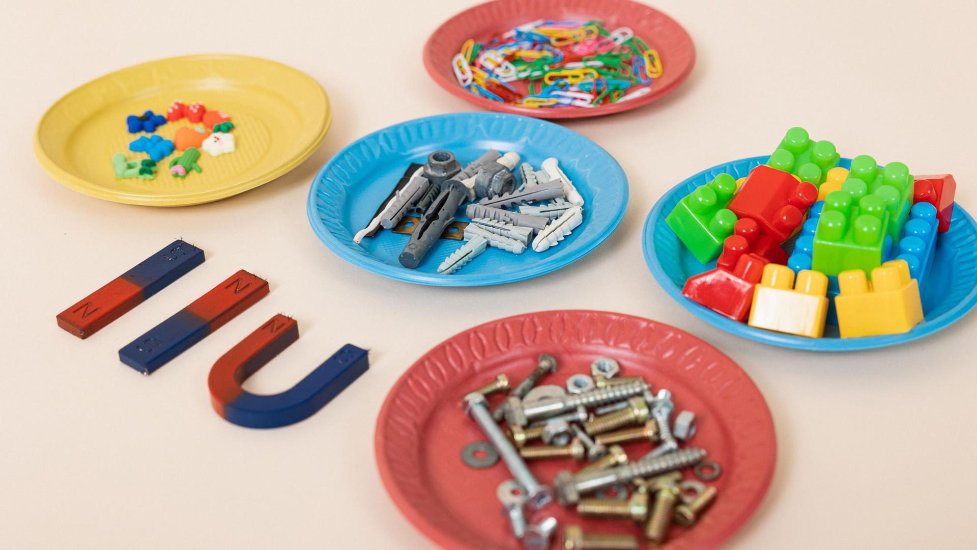 3 Magnete liegen auf einem Tisch, daneben diverse bunte Teller mit unterschiedlichen Materialien 3 Magnete liegen auf einem Tisch, daneben diverse bunte Teller mit unterschiedlichen Materialien