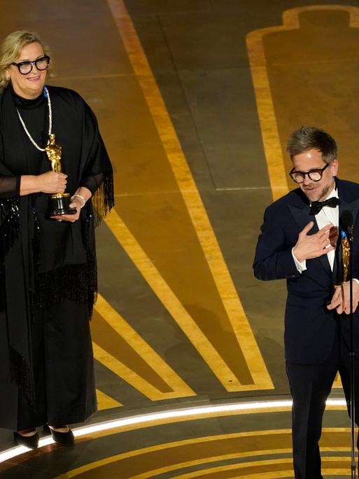 Ernestine Hipper im schwarzen Kleid und Christian M. Goldbeck im Smoking halten jeweils eine goldene Oscar-Statue in Händen.