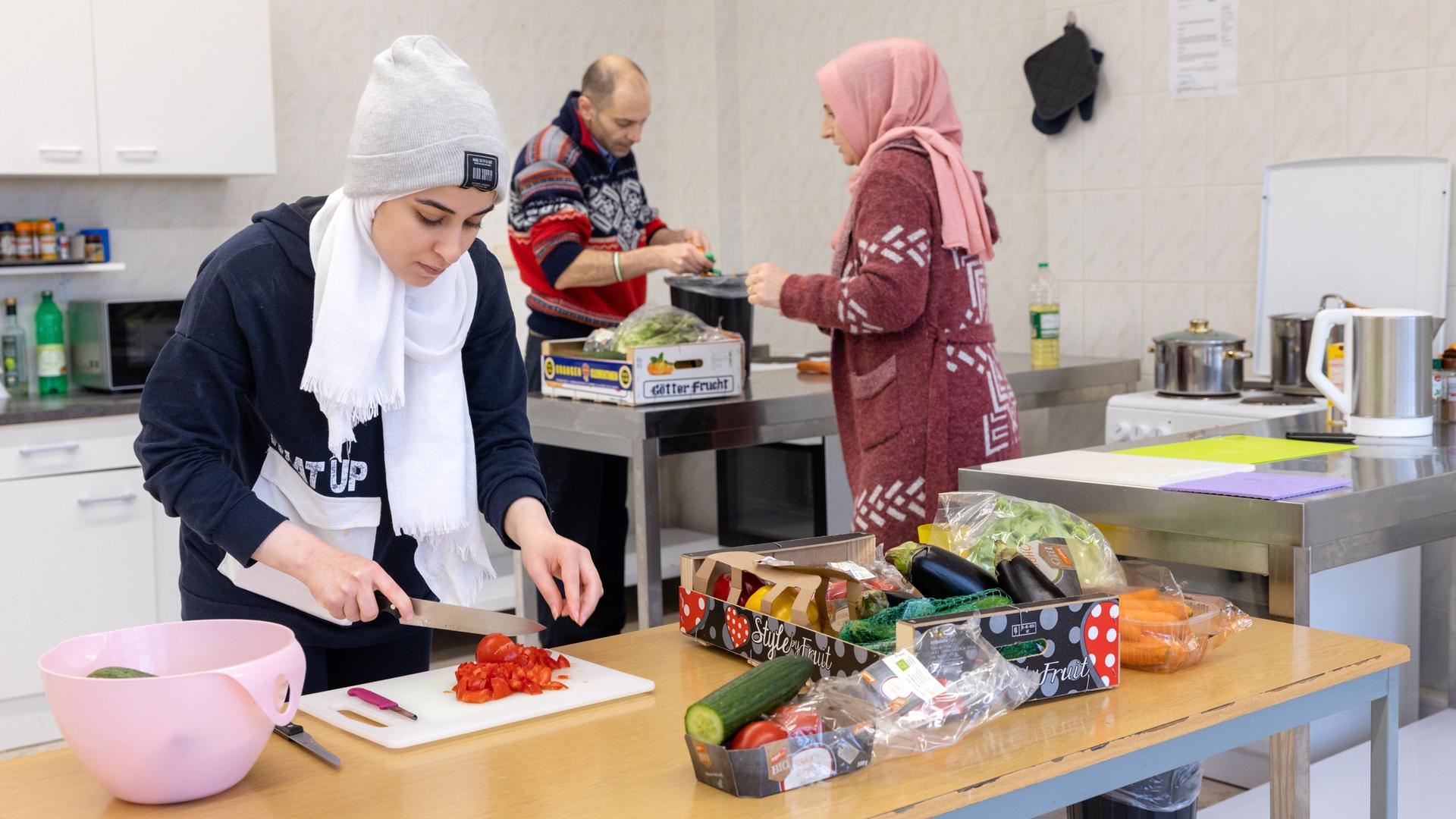 Zwei Frauen mit Kopftüchern und ein Mann stehen in einer Küche und berieten eine Mahlzeit vor. 