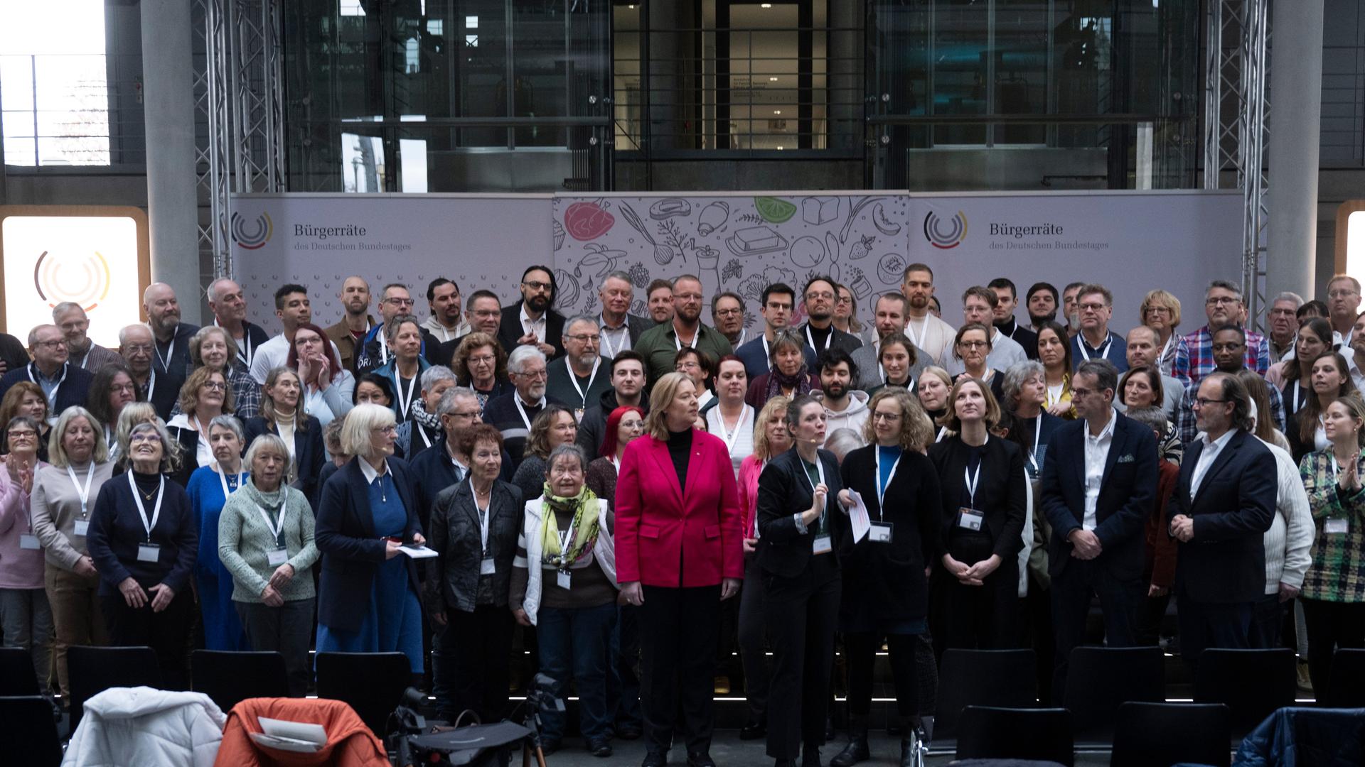Die Mitglieder des Bürgerrats stehen aufgereiht im Bundestag, vorne in der Mitte steht Bundestagspräsidentin Bärbel Bas. Sie trägt ein pinkfarbenes Sakko.
