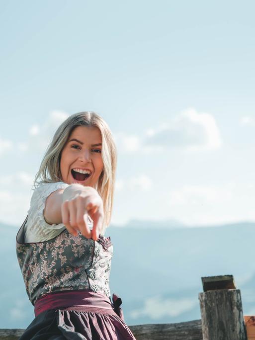 Eine junge Frau in Dirndl steht vor einer Bergkette und lacht in die Kamera.