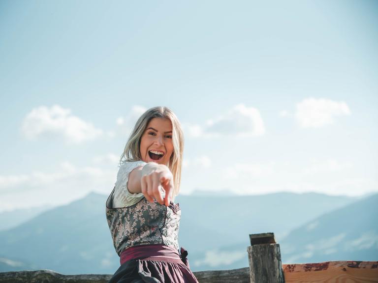 Eine junge Frau in Dirndl steht vor einer Bergkette und lacht in die Kamera.