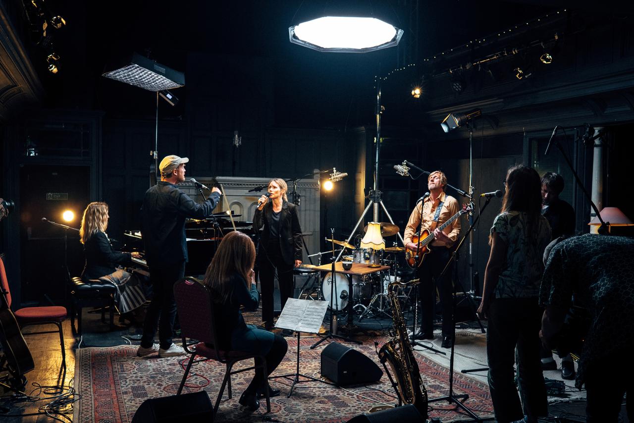 In einem gemütlichen Studioraum stehen und sitzen Musikerinnen und Musiker um James Yorkston und spielen während einer Aufnahme.
