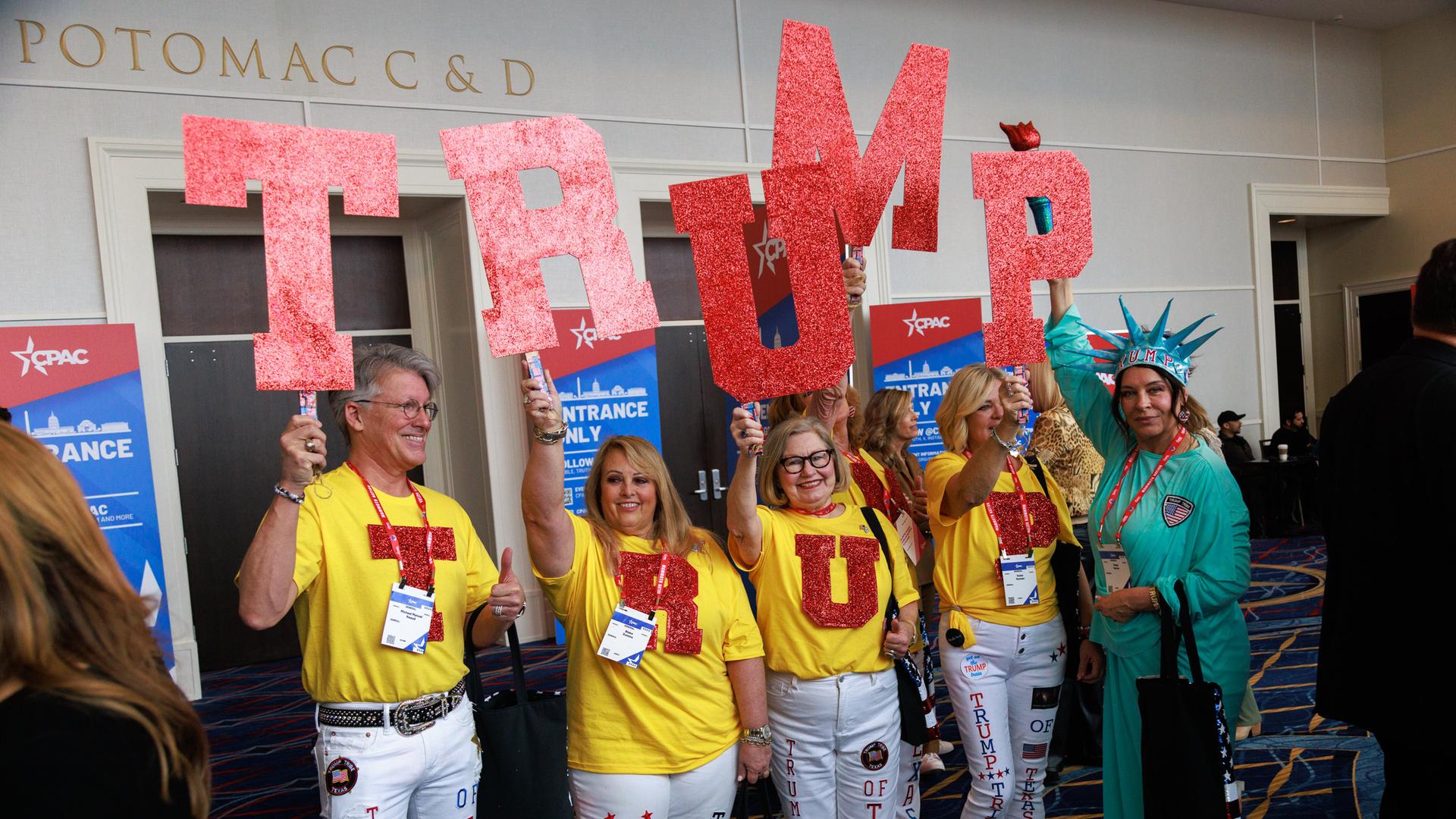 Ein Gruppe von Menschen in gelben T-Shirts steht aufgereiht nebeneinander. Sie halten rote Buchstaben in die Höhe, die den Namen "Trump" ergeben