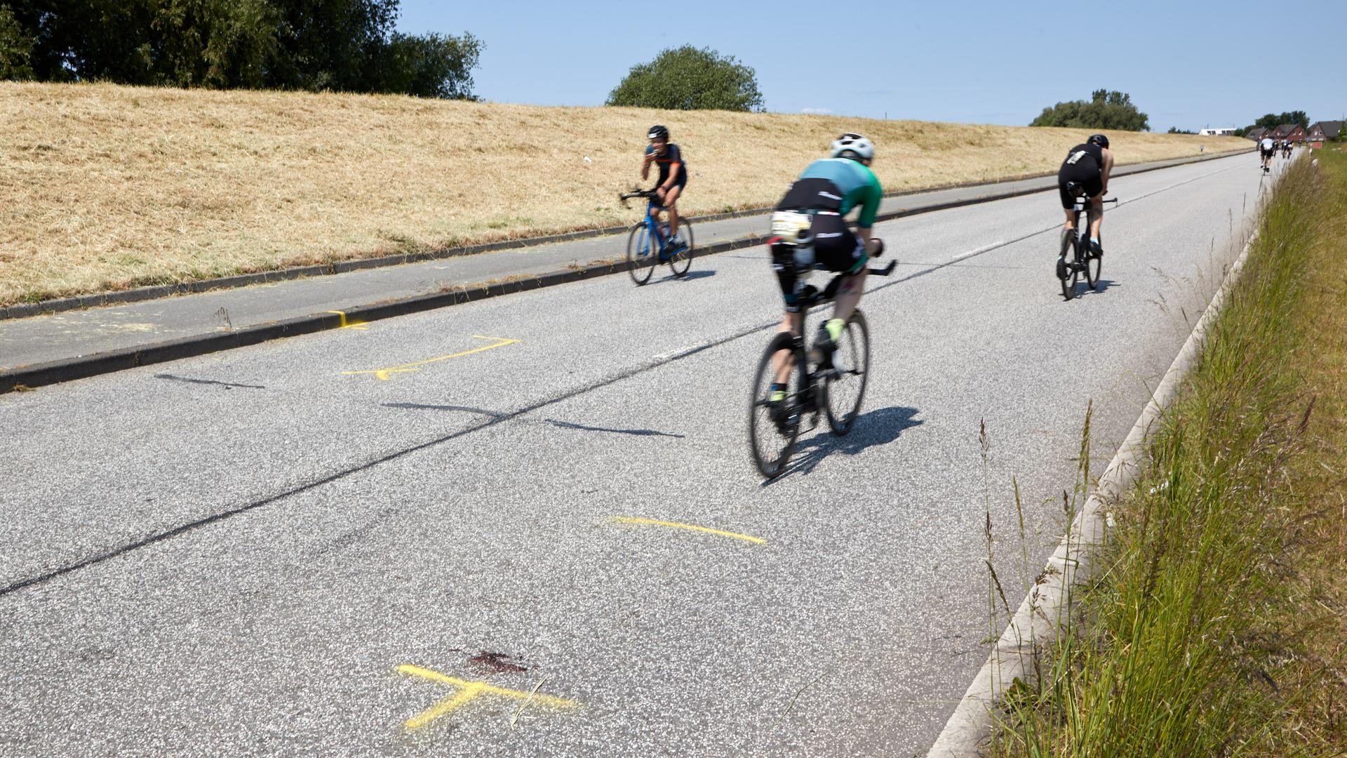 Triathleten fahren auf dem Rad an der Unglückstelle auf dem Gaueter Hauptdeich vorbei. Die Stelle ist mit gelber Farbe markiert.