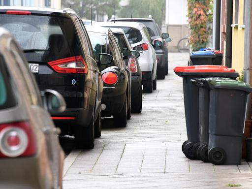 Autos auf dem Gehweg, der schon mit Mülltonnen zugestellt ist. 