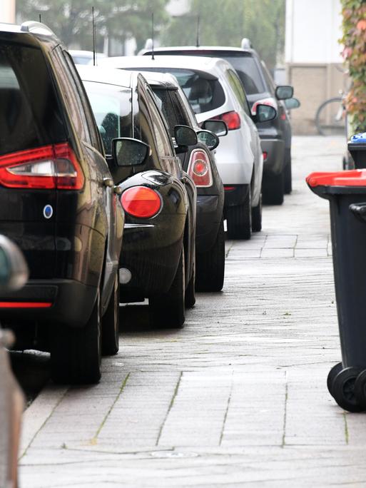 Autos auf dem Gehweg, der schon mit Mülltonnen zugestellt ist. 