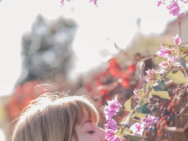 Eine Frau mit Jacke und Schal riecht draußen an einer Wand an Blumen