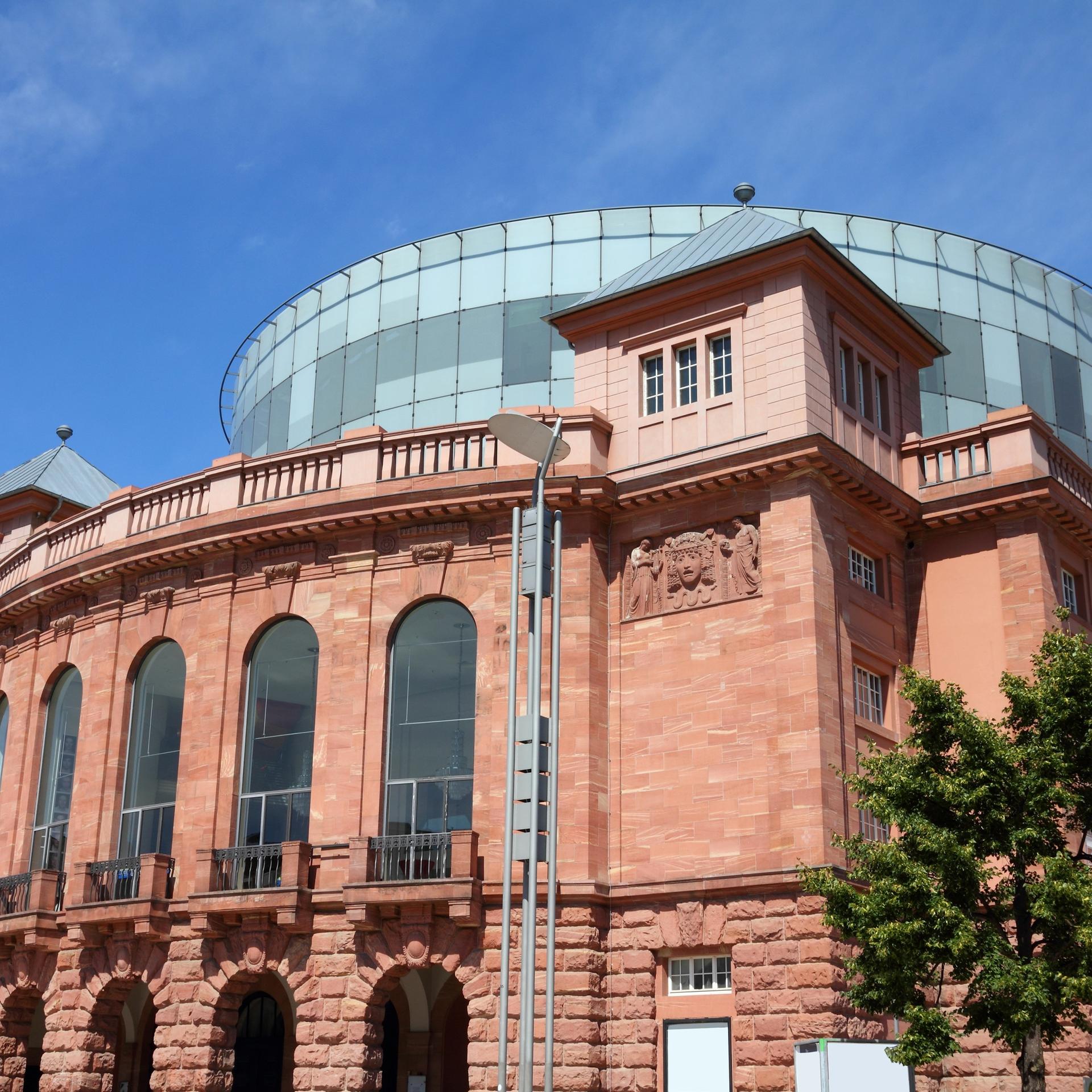 Das Foto zeigt das Staatstheater in Mainz, ein älteres Gebäude verkleidet mit roten Ziegeln und einem Glasdach. 