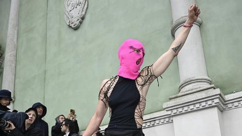 Eine Frau mit knallpinker Strickmaske steht auf dem steinernden Treppengeländer vor dem russischen Pavillon auf der Biennale in Venedig, reckt ihre Faust in die Luft und schreit.