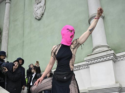 Eine Frau mit knallpinker Strickmaske steht auf dem steinernden Treppengeländer vor dem russischen Pavillon auf der Biennale in Venedig, reckt ihre Faust in die Luft und schreit.