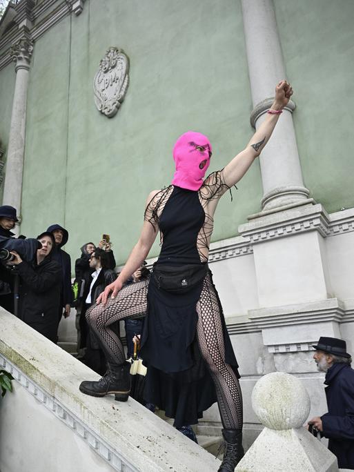 Eine Frau mit knallpinker Strickmaske steht auf dem steinernden Treppengeländer vor dem russischen Pavillon auf der Biennale in Venedig, reckt ihre Faust in die Luft und schreit.