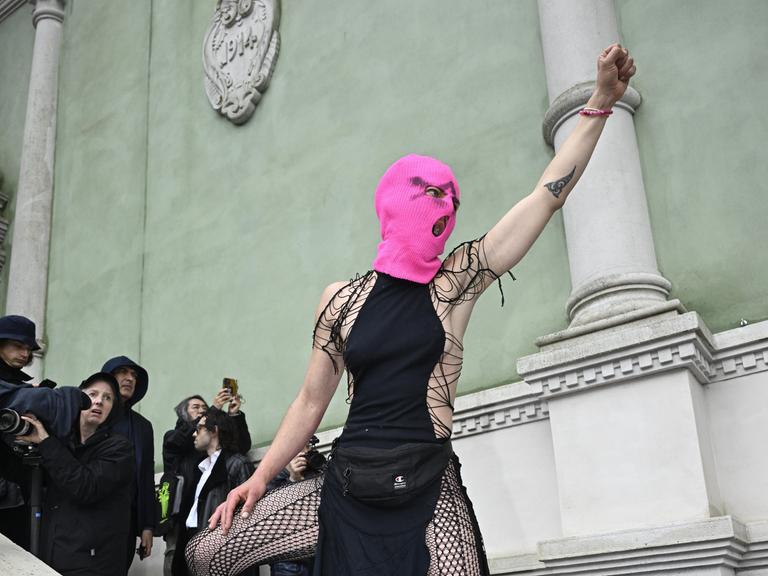 Eine Frau mit knallpinker Strickmaske steht auf dem steinernden Treppengeländer vor dem russischen Pavillon auf der Biennale in Venedig, reckt ihre Faust in die Luft und schreit.