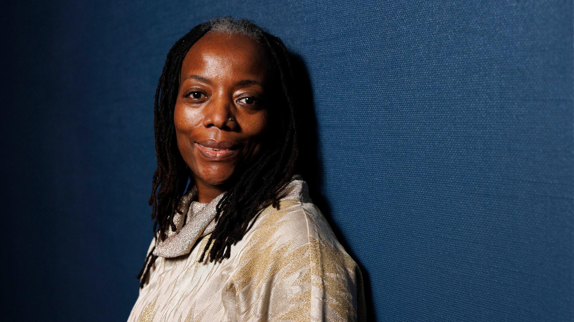 Porträtaufnahme der simbabwischen Autorin und Filmemacherin Tsitsi Dangarembga. Sie trägt ein helles Oberteil, ihre Haare sind zu vielen Zöpfen geflochten und sie steht vor einem graublauen Hintergrund.