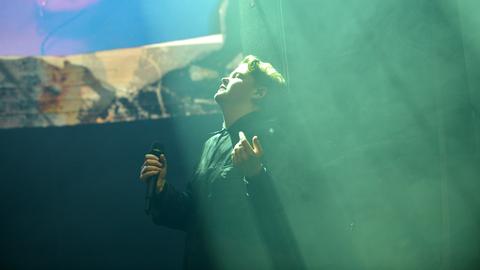Kae Tempest steht bei einem Auftritt auf der Bühne, hat die Augen geschlossen und den Kopf nach oben gedreht, in Richtung eines grünen Lichtstrahls von der Decke.