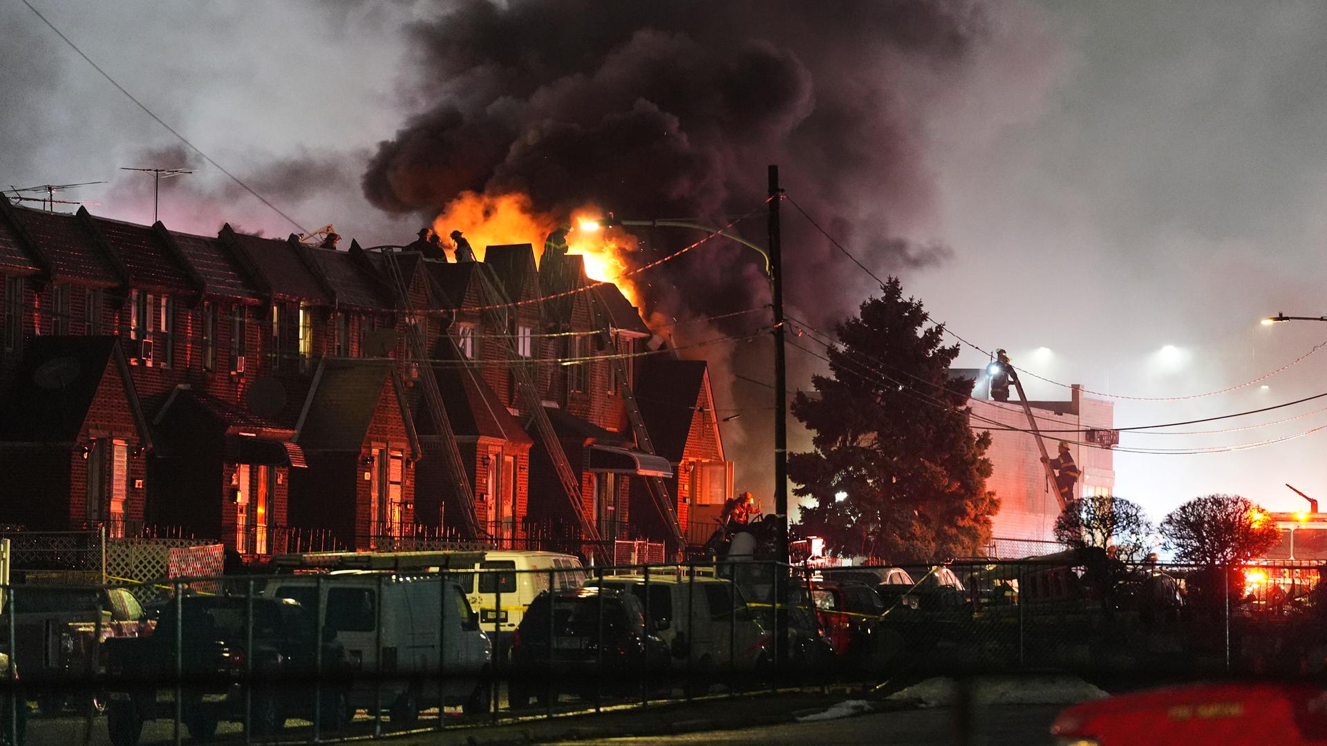 Blick auf eine Häuserreihe, hinter der hohe Flammen in die Dunkelheit lodern. 