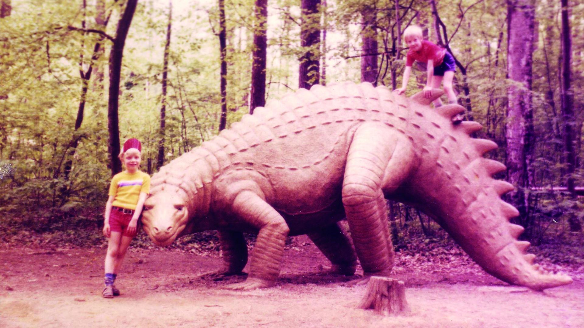 Zwei Kinder posieren neben dem lebensgroßen Modell eines Dinosauriers in einem Freizeitpark.