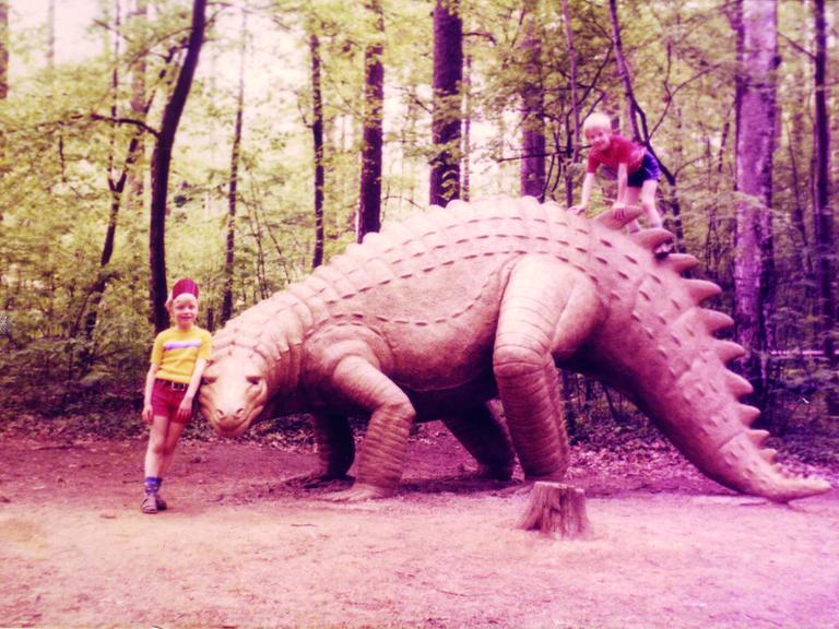 Zwei Kinder posieren neben dem lebensgroßen Modell eines Dinosauriers in einem Freizeitpark.