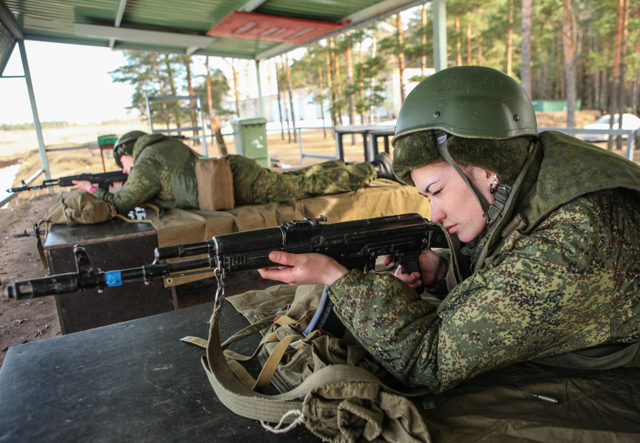 Zwei Frauen in Tarnuniform und mit Helm liegen auf einem Militärgelände unter einem Dach auf Holzkisten und zielen mit ihren Waffen nach vorne. 