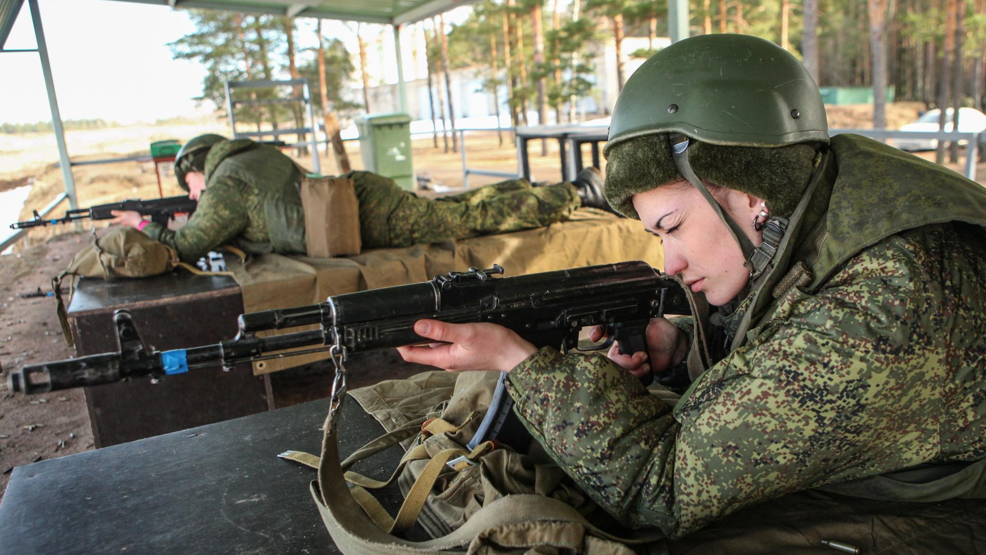 Zwei Frauen in Tarnuniform und mit Helm liegen auf einem Militärgelände unter einem Dach auf Holzkisten und zielen mit ihren Waffen nach vorne.