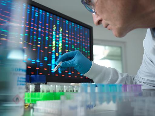 Ein Wissenschaftler hält ein Reagenzglas vor einen Bildschirm mit Gensequenzen.