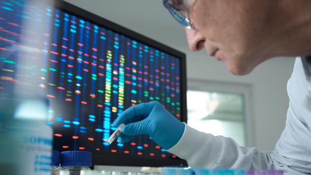 Ein Wissenschaftler hält ein Reagenzglas vor einen Bildschirm mit Gensequenzen.