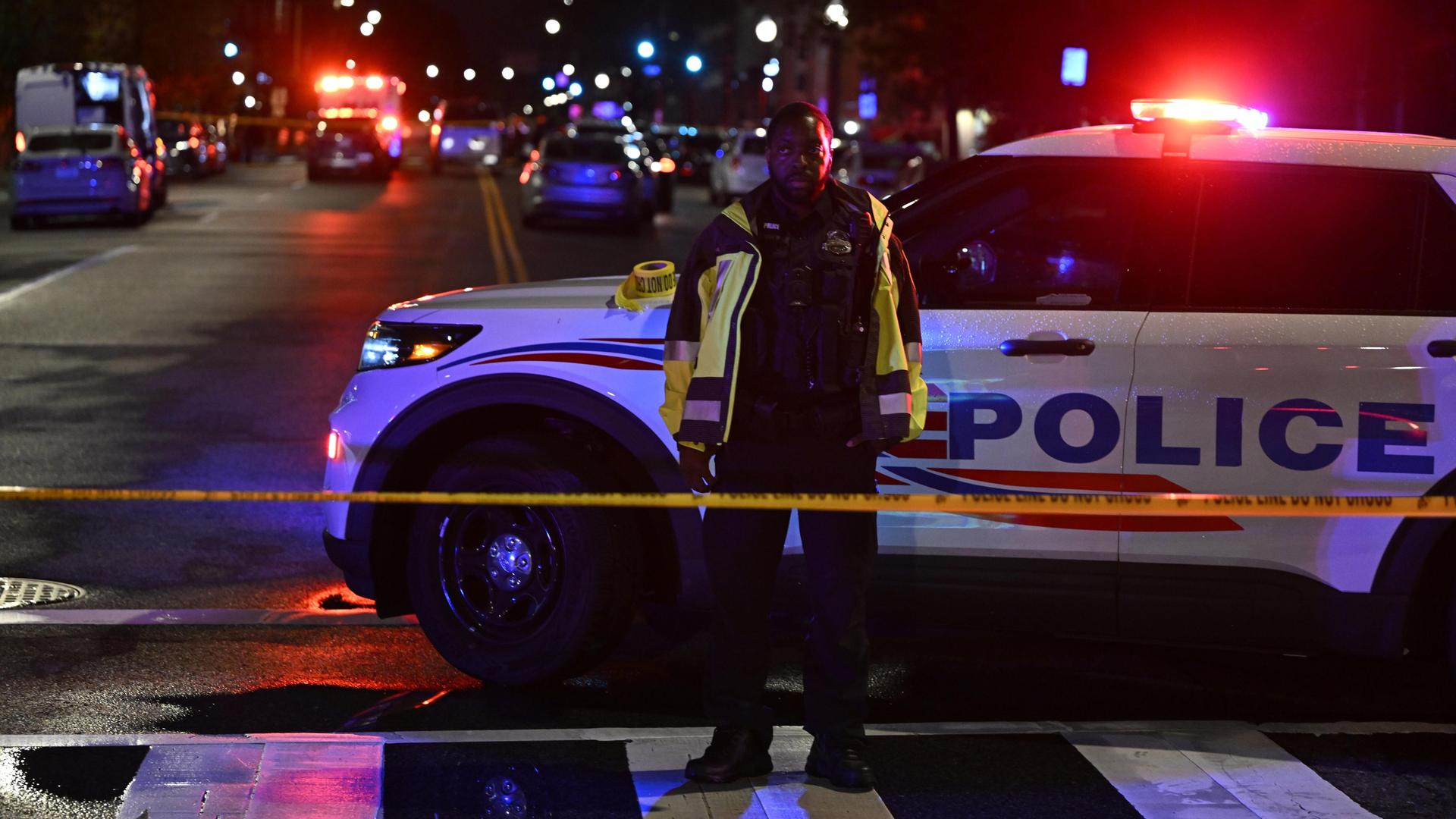 Ein US-Polizist steht vor seinem Polizeiwagen in Washington, im Hintergrund ist ein Tatort abgesperrt.