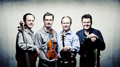 Die vier Musiker stehen vor einem hellen Hintergrund eng beieinander und haben zum Teil ihre Instrumente dabei. Die vier Musiker stehen vor einem hellen Hintergrund eng beieinander und haben zum Teil ihre Instrumente dabei.