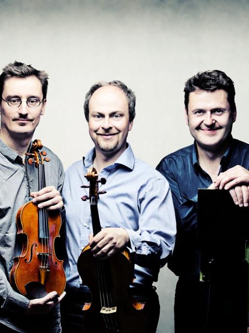 Die vier Musiker stehen vor einem hellen Hintergrund eng beieinander und haben zum Teil ihre Instrumente dabei.