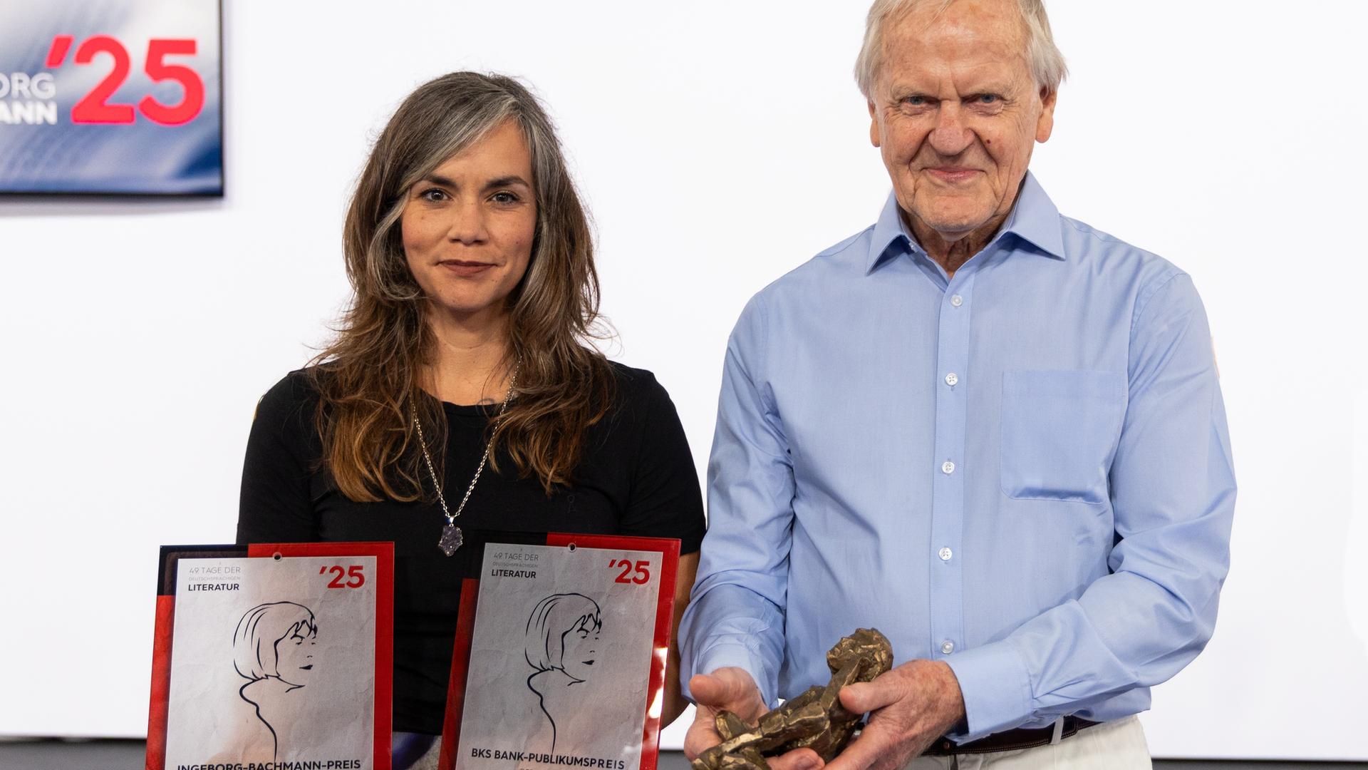 Natascha Gangl hält ihren Bachmannpreis in der Hand und lächelt. Neben ihr steht Heinz Bachmann, der Bruder von Ingeborg Bachmann. Auch er lächelt.