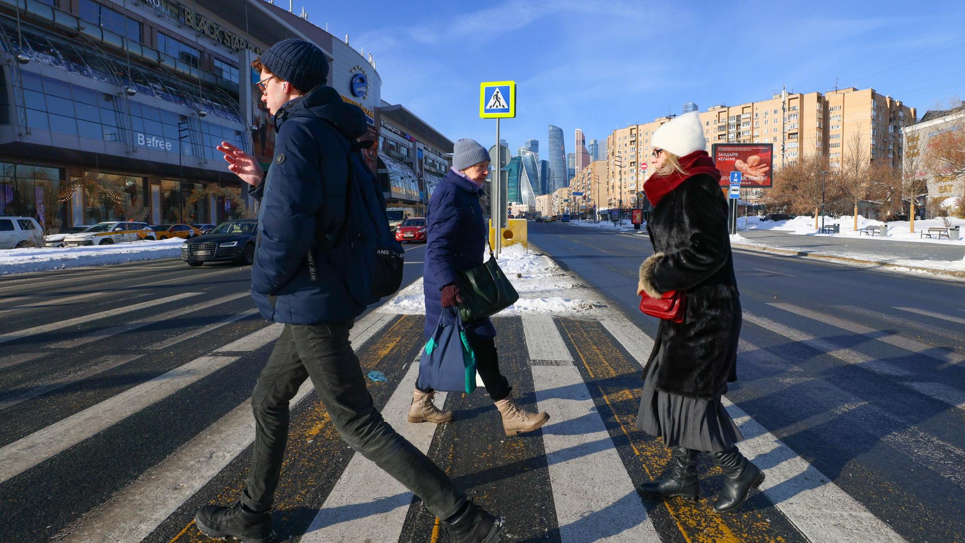 Ein junger Mann läuft rauchend über den Zebrastreifen einer großen Straße, hinter ihm eine Frau mit Brille und Handtasche. In die andere Richtung läuft eine ältere Frau mit zwei Umhängetaschen.