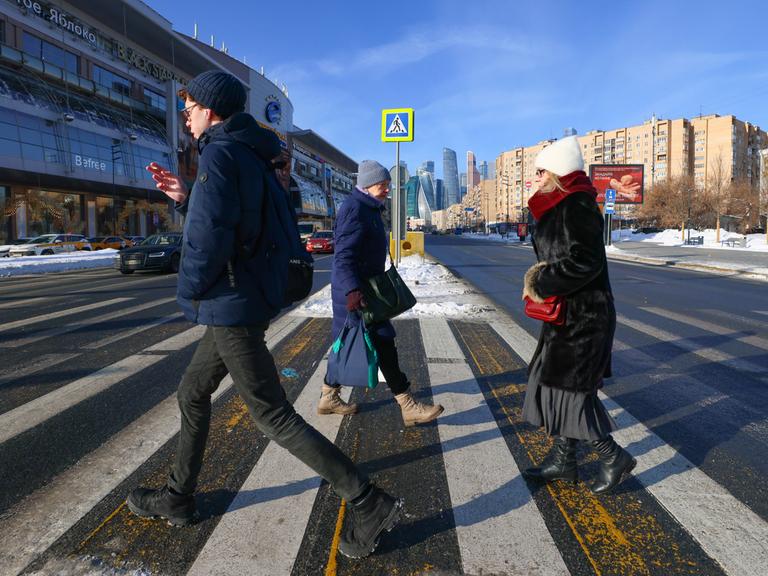Ein junger Mann läuft rauchend über den Zebrastreifen einer großen Straße, hinter ihm eine Frau mit Brille und Handtasche. In die andere Richtung läuft eine ältere Frau mit zwei Umhängetaschen.