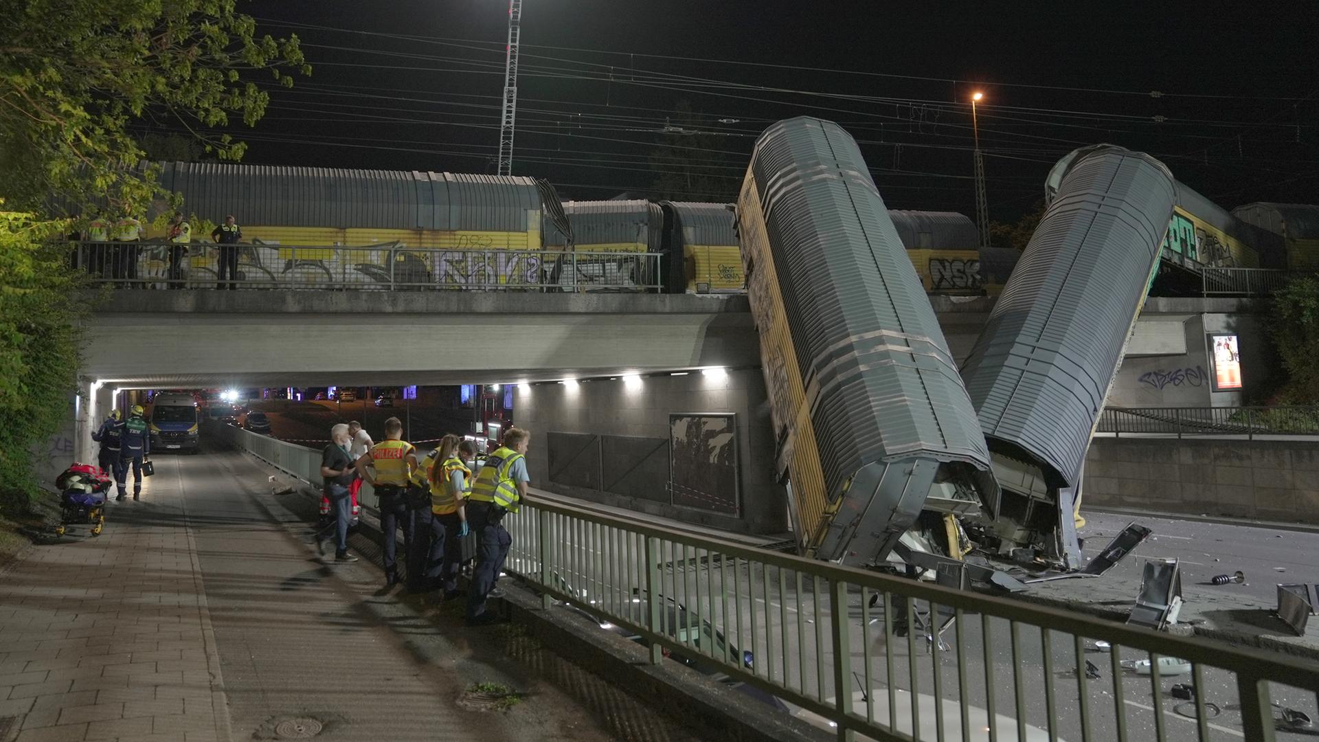 Zwei Waggons eines Güterzugs liegen schräg senkrecht auf einer Straße, nachdem sie von einer Brücke gestürzt sind. Im Vordergrund sind Einsatzkräfte zu sehen.