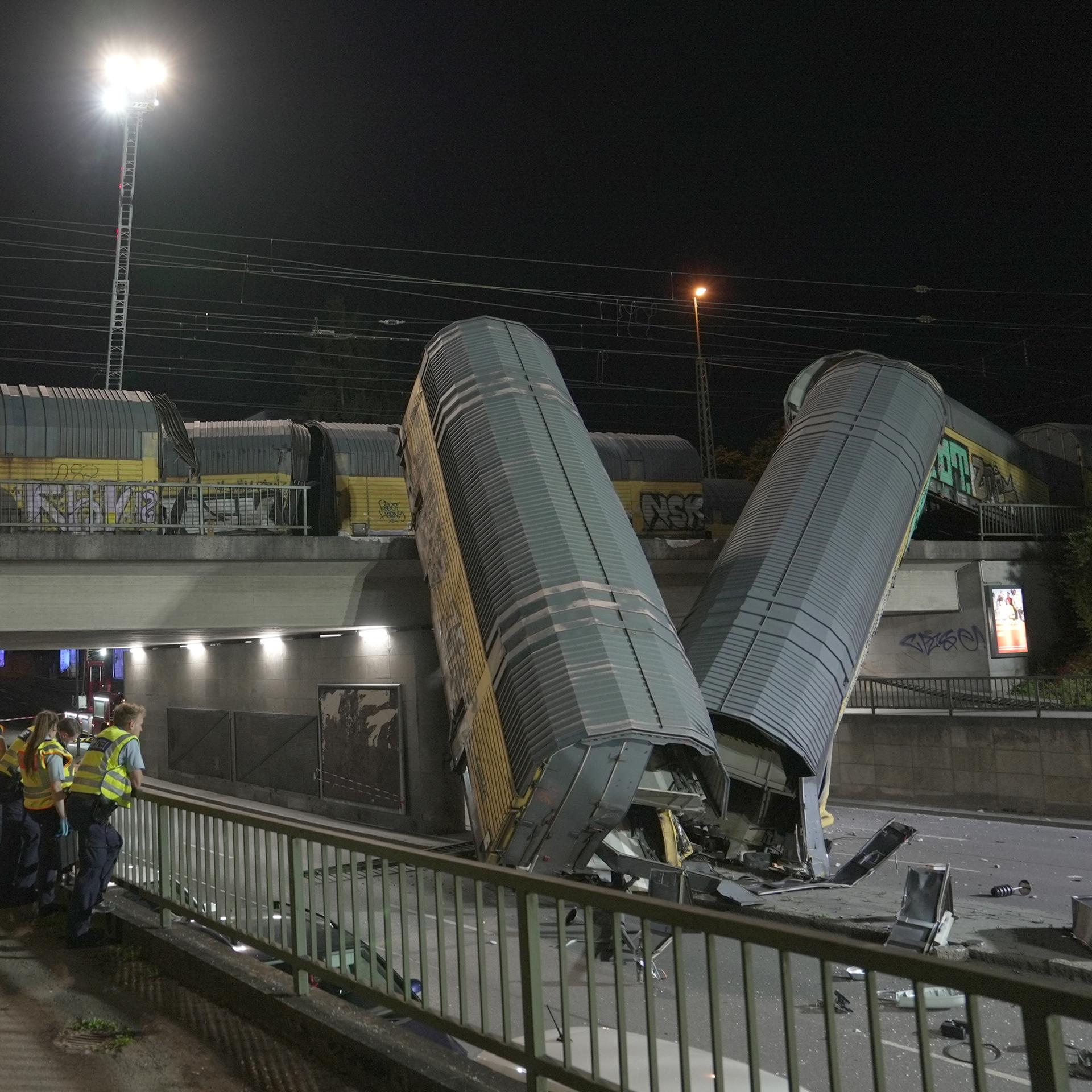 Zwei Waggons eines Güterzugs liegen schräg senkrecht auf einer Straße, nachdem sie von einer Brücke gestürzt sind. Im Vordergrund sind Einsatzkräfte zu sehen.