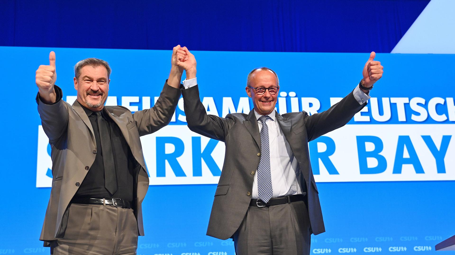 Markus Söder (links) steht beim CSU-Parteitag auf der Bühne neben Friedrich Merz: Söder und Merz halten die linke bzw. rechte Hand des anderen und halten diese hoch, den anderen Arm strecken sie nach vorn und halten den Daumen hoch.