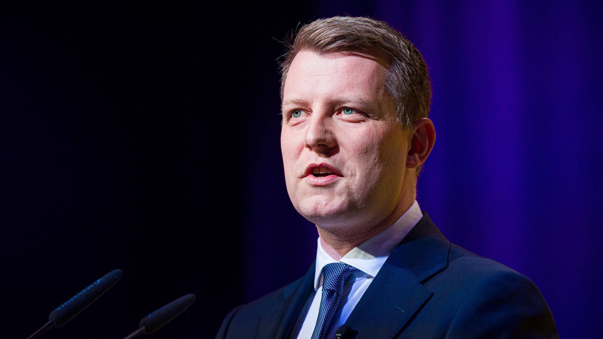 Henning Höne mit Jackett und Krawatte spricht auf einer Bühne in ein Mikrofon. De Hintergrund ist dunkel. Henning Höne mit Jackett und Krawatte spricht auf einer Bühne in ein Mikrofon. De Hintergrund ist dunkel.