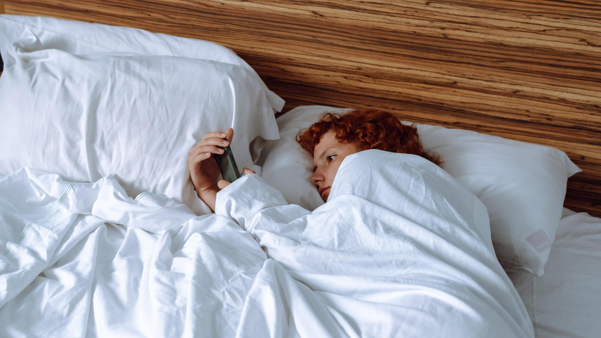Eine rothaarige Person liegt im Bett auf der Seite, die Bettdecke bis ins Gesicht gezogen, und schaut auf das Smartphone, das sie in der Hand hält.