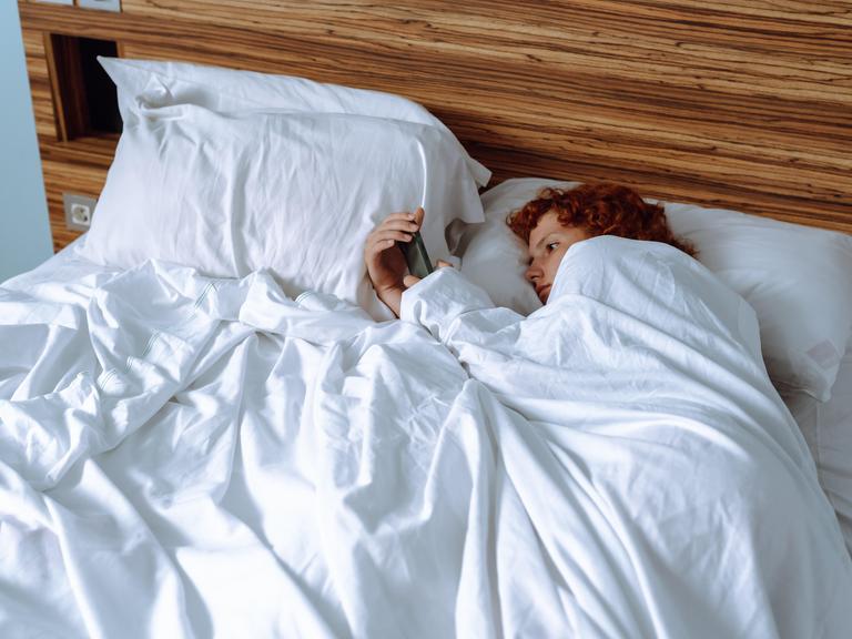 Eine rothaarige Person liegt im Bett auf der Seite, die Bettdecke bis ins Gesicht gezogen, und schaut auf das Smartphone, das sie in der Hand hält.