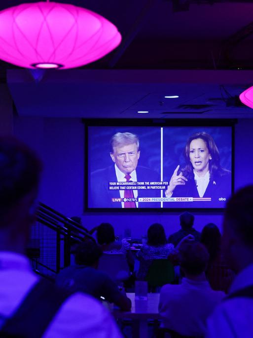 Menschen in einer Bar verfolgen auf einem großen TV-Bildschirm die Debatte zwischen Kamala Harris und Donald Trump.