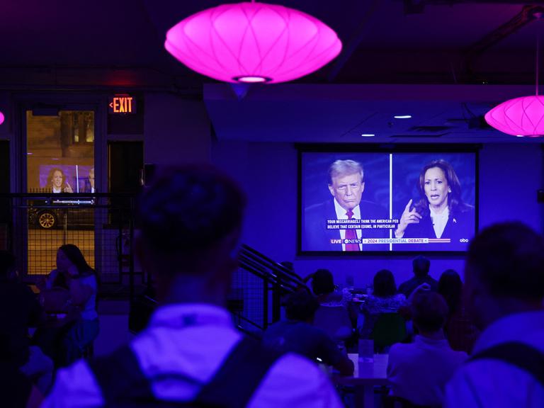 Menschen in einer Bar verfolgen auf einem großen TV-Bildschirm die Debatte zwischen Kamala Harris und Donald Trump.