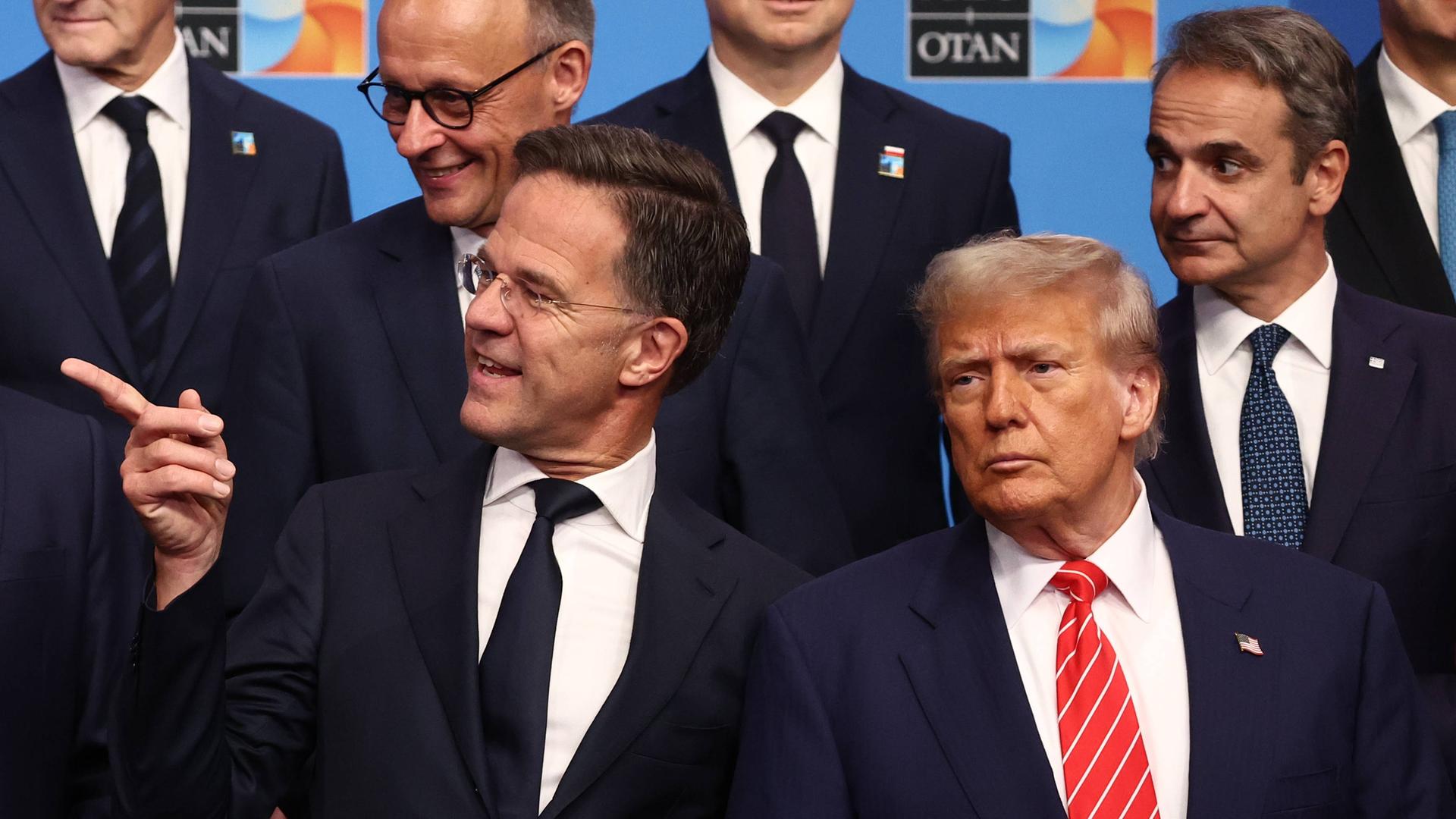 Beim NATO-Gipfel in Den Haag stehen Friedrich Merz, Mark Rutte und Donald Trump mit weiteren Politikern vor einer blauen Fotowand. Rutte zeigt mit dem Finger nach links. Beim NATO-Gipfel in Den Haag stehen Friedrich Merz, Mark Rutte und Donald Trump mit weiteren Politikern vor einer blauen Fotowand. Rutte zeigt mit dem Finger nach links.