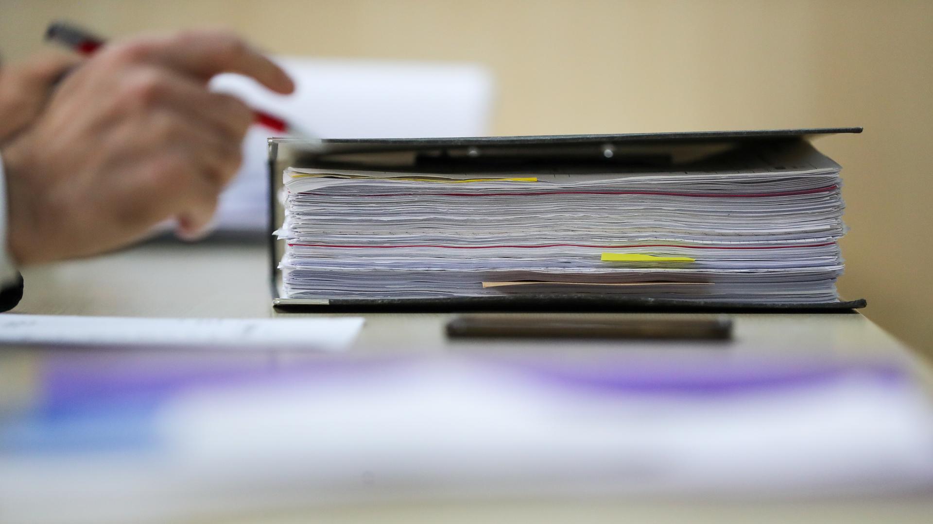 Ein Aktenordner liegt auf einem Tisch, im Hintergrund in der Unschärfe ist die Hand einer Person zu sehen, die einen Stift hält. 