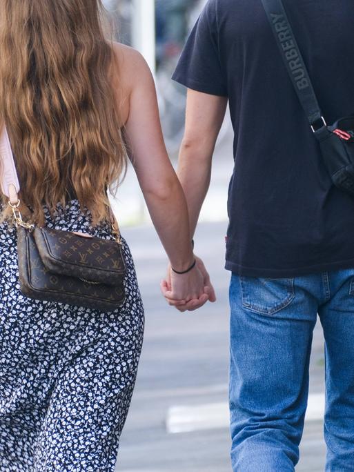 Ein junges Paar fotografiert von hinten Händchenhaltend beim Spaziergang durch eine Stadt. Sie trägt eine Louis Vuitton Tasche und hat lange Haare.